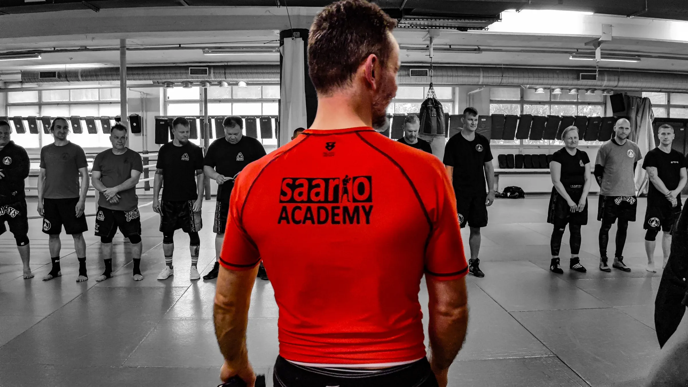 A Saario Academy instructor in the red Academy shirt, seen from behind, facing a lined-up class in a training hall
